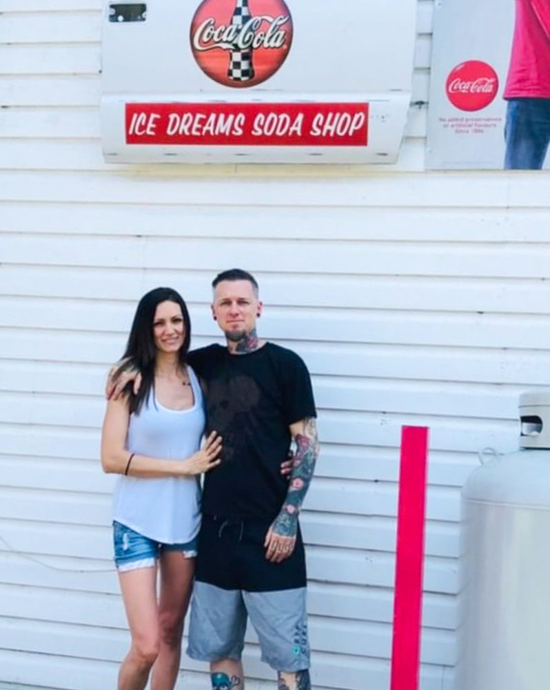 A dark-haired woman and a tattooed man stand under a sign that reads "Ice Dreams Soda Shop".