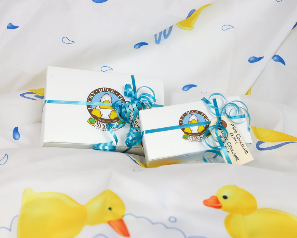 2 gift wrapped boxes of Lucky Duck Fudge, decorate with a blue, curled ribbon and gift tags with fudge flavours. Set on a white background with colourful rubber ducks and water droplets.