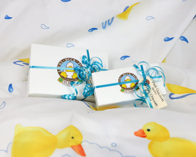 2 gift wrapped boxes of Lucky Duck Fudge, decorate with a blue, curled ribbon and gift tags with fudge flavours. Set on a white background with colourful rubber ducks and water droplets.