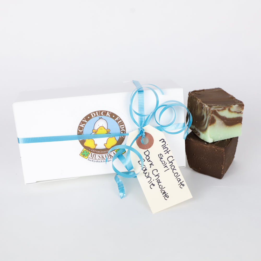 A small white box of gift wrapped fudge, decorated with a blue, curled ribbon and a tag. A stack of mint chocolate swirl fudge and dark chocolate brownie fudge sit next to the box.